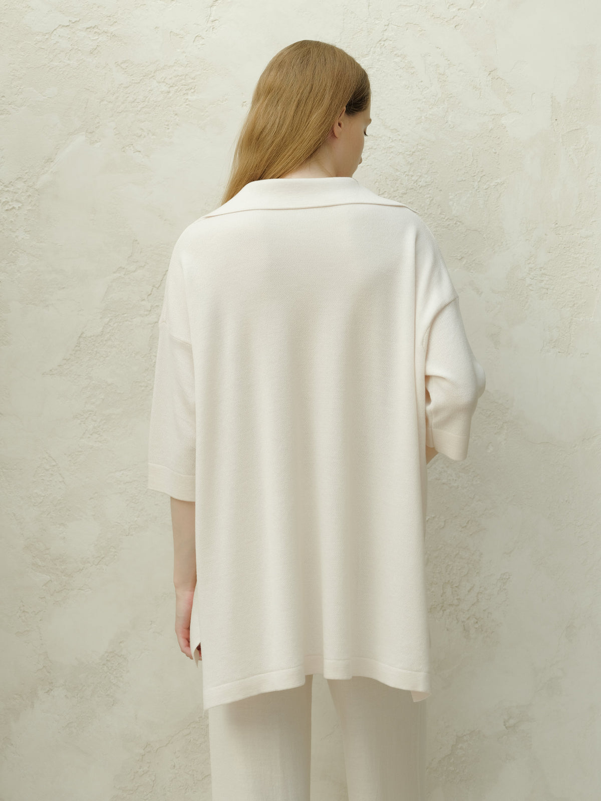 Vista posterior de una modelo con Merino Wool Maxi Polo crema pálido de caída suelta frente a una pared texturizada