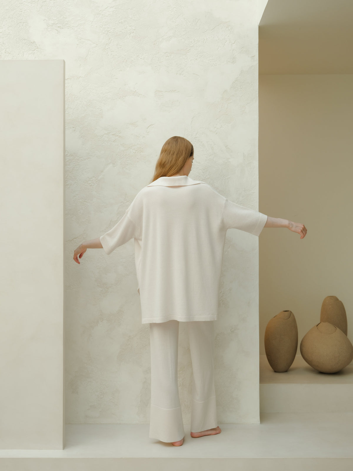 Vista posterior de una modelo con maxi polo oversize blanco de lana merino frente a una pared clara texturizada