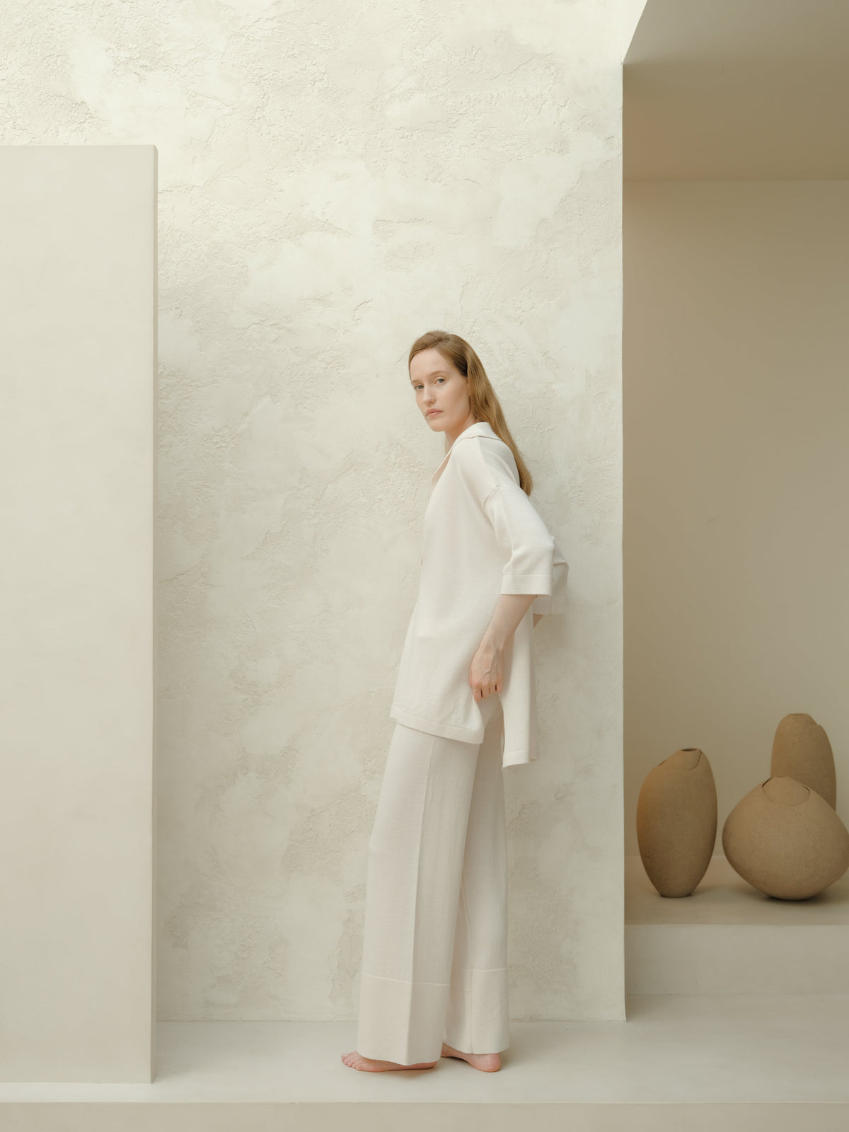 Modelo con a white Maxi polo de lana merina extrafina and pantalones de pierna ancha a juego stands barefoot against a pale textured wall.