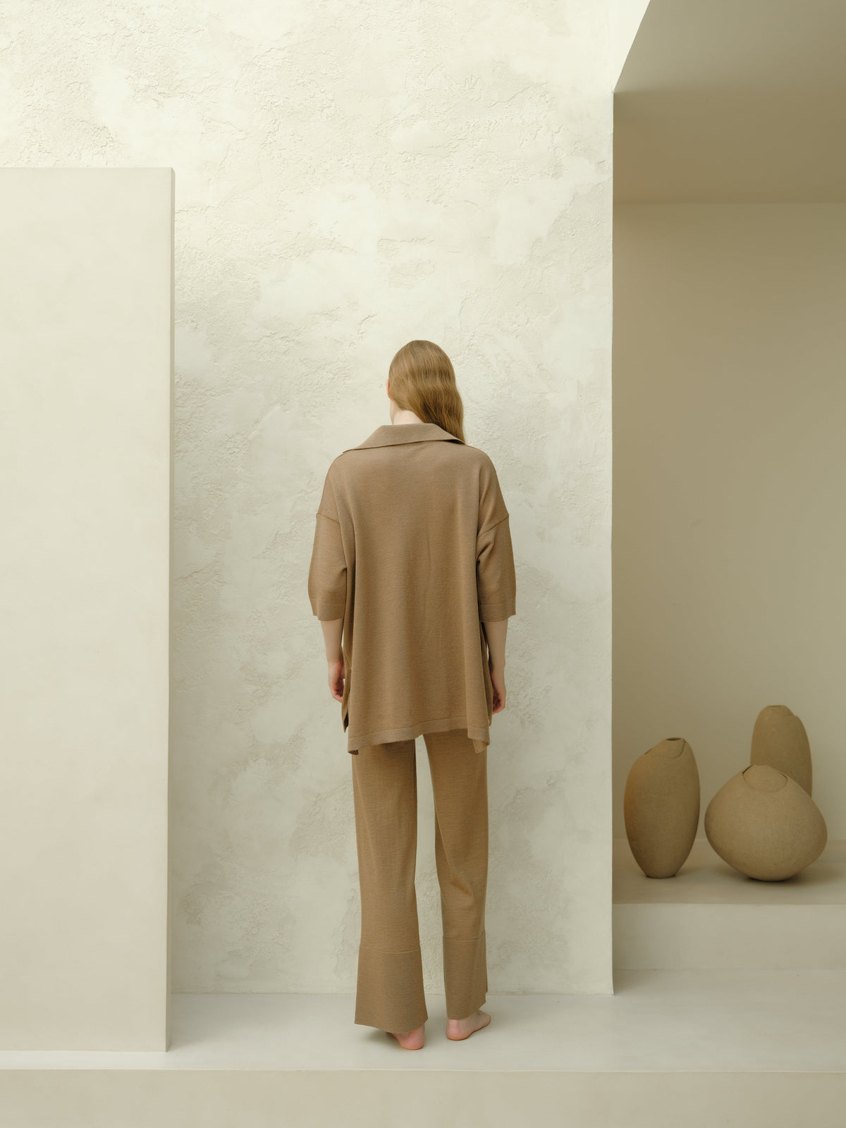 Back view of a woman in camel brown merino wool maxi polo set de pie y descalza in a minimalist studio.
