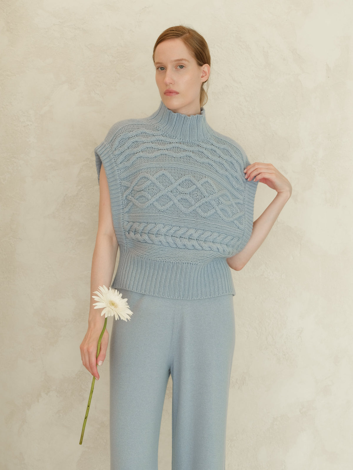 Model trägt blue cashmere mix Maxi-Zopfstricktunika with a high ribbed neck and sleeveless silhouette against a textured wall.