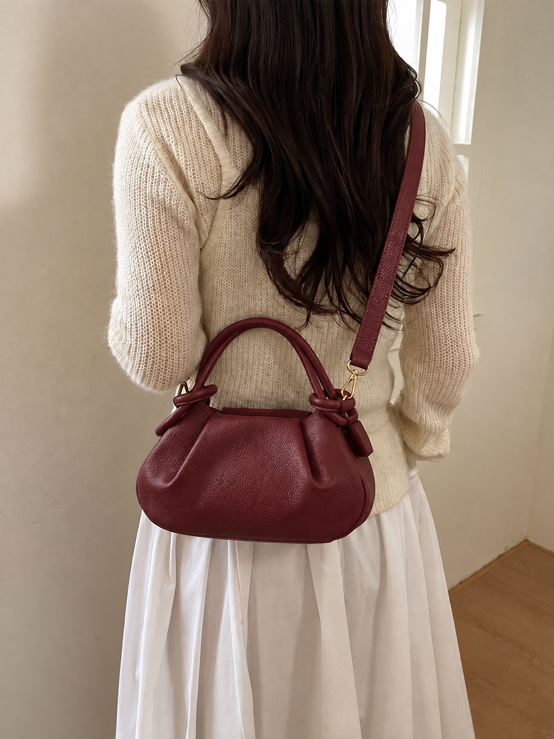 Mujer con jersey de punto crema y falda blanca lleva un pequeño bolso de piel burdeos con asa superior y correa de hombro, vista de espaldas en un interior.