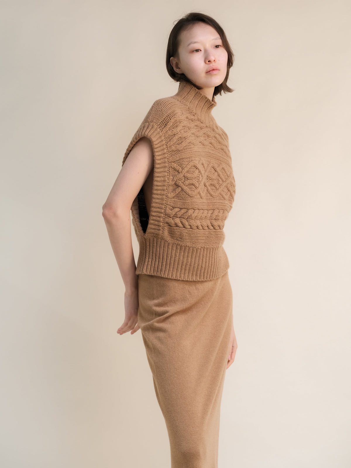 Profile view of a Model trägt a tan cashmere mix Maxi-Zopfstricktunika with a high ribbed neck and bold knit patterns.
