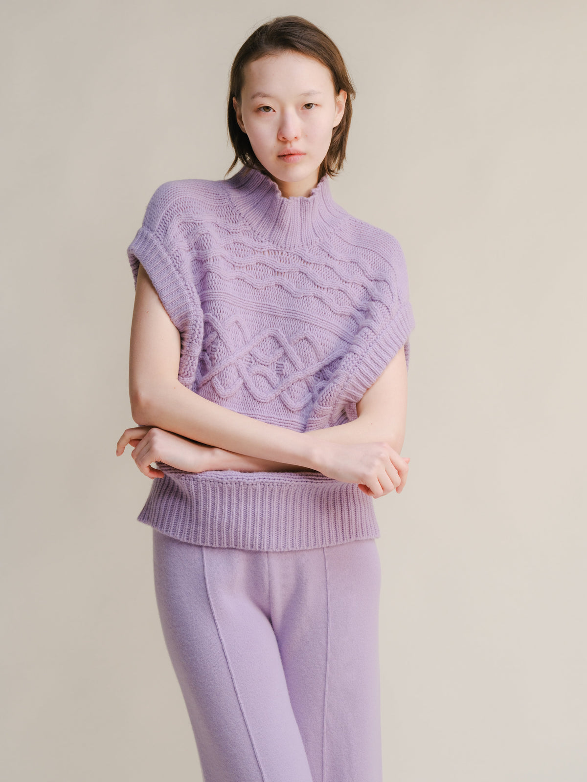 Flieder chunky knit sleeveless tunic with a hoher Kragen and cable pattern worn with matching lilac pants.