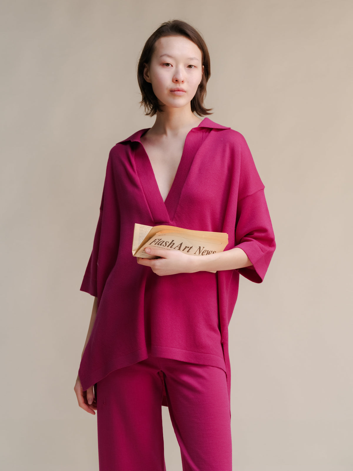 Rosa intenso merino wool maxi polo worn by a model with a deep V neckline, holding a folded newspaper