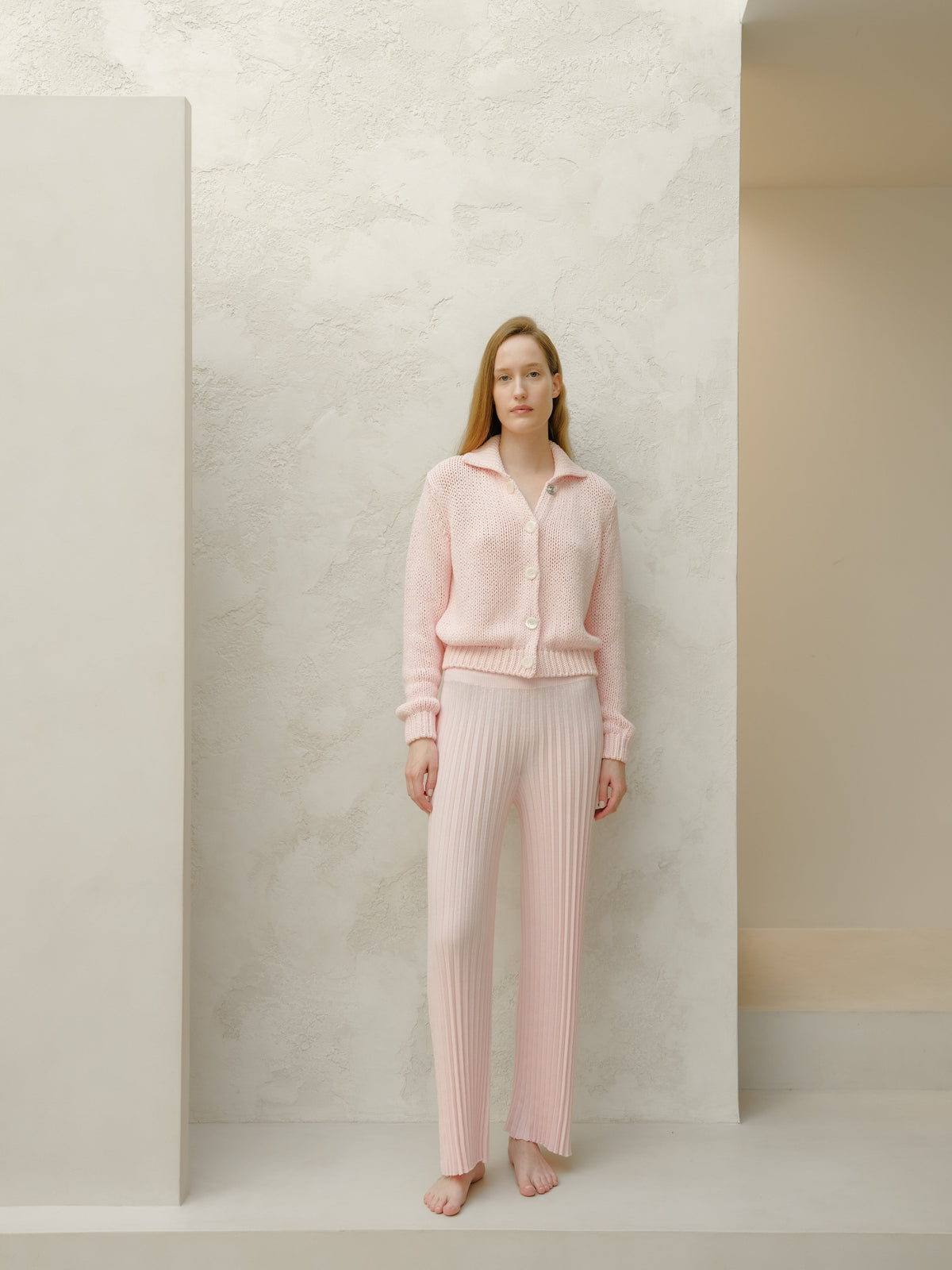 Modella con pale rosa knitted cardigan e matching ribbed pants in piedi a piedi nudi against a texturosso pale wall.