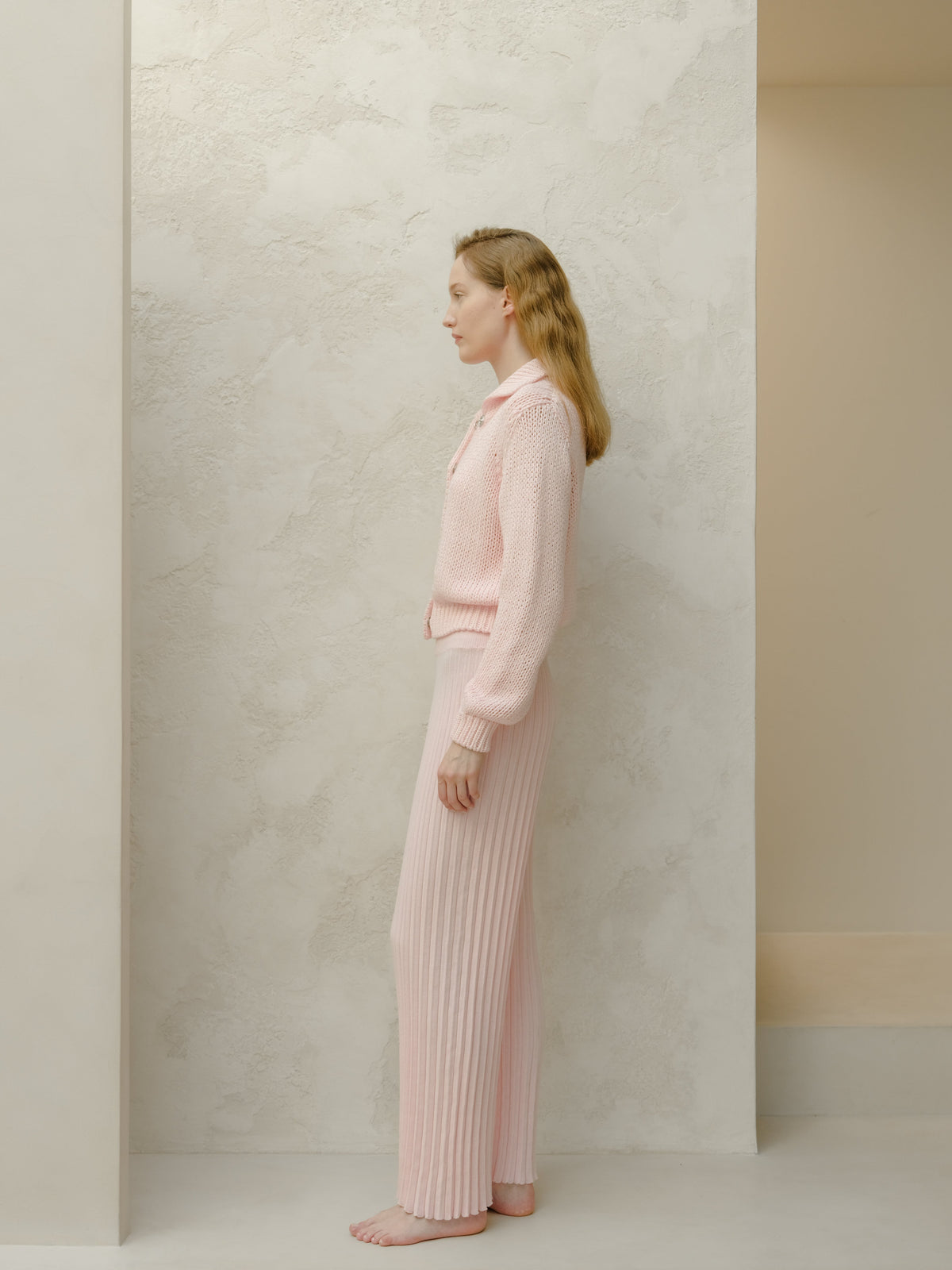 Vista laterale of a model in a pale rosa knit set con a sweater e ribbed pantaloni a gamba larga against a texturosso pale wall