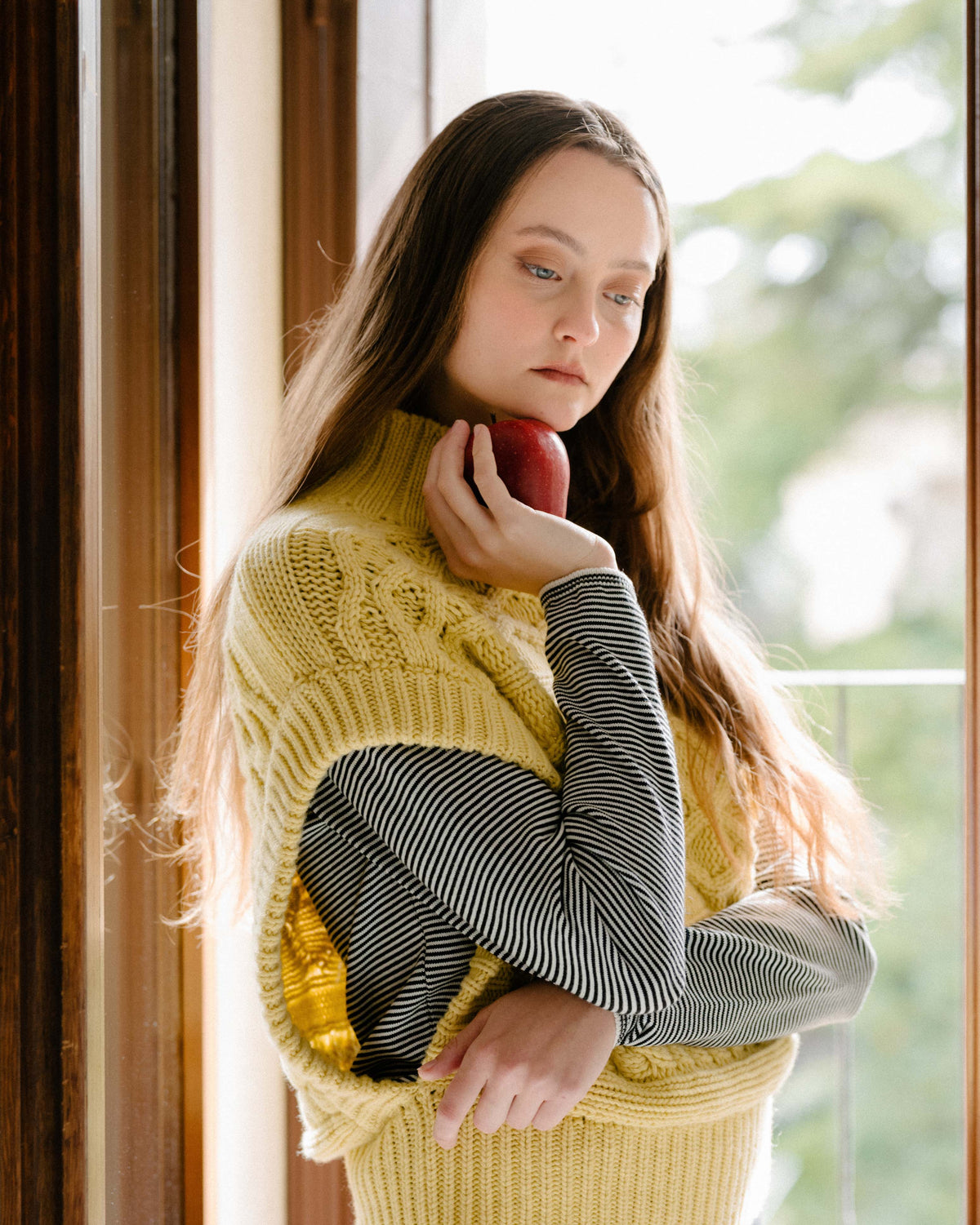 Weibliches Model trägt a cream cashmere mix Maxi-Zopfstricktunika over a striped top, holding a red apple near her face.