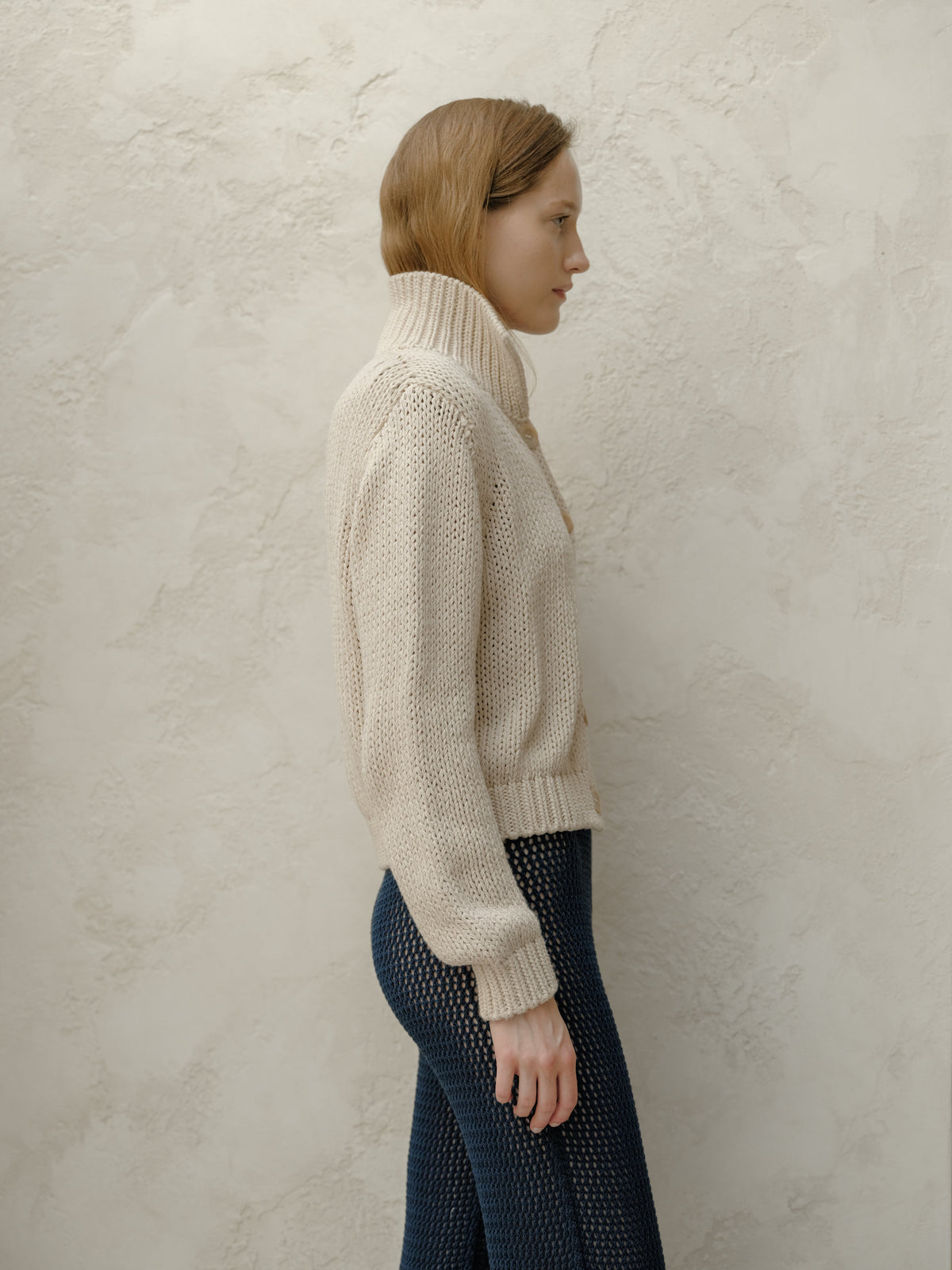 Vista laterale of model in a beige knit bomber jacket con a high collar abbinata a blu navy openwork pants