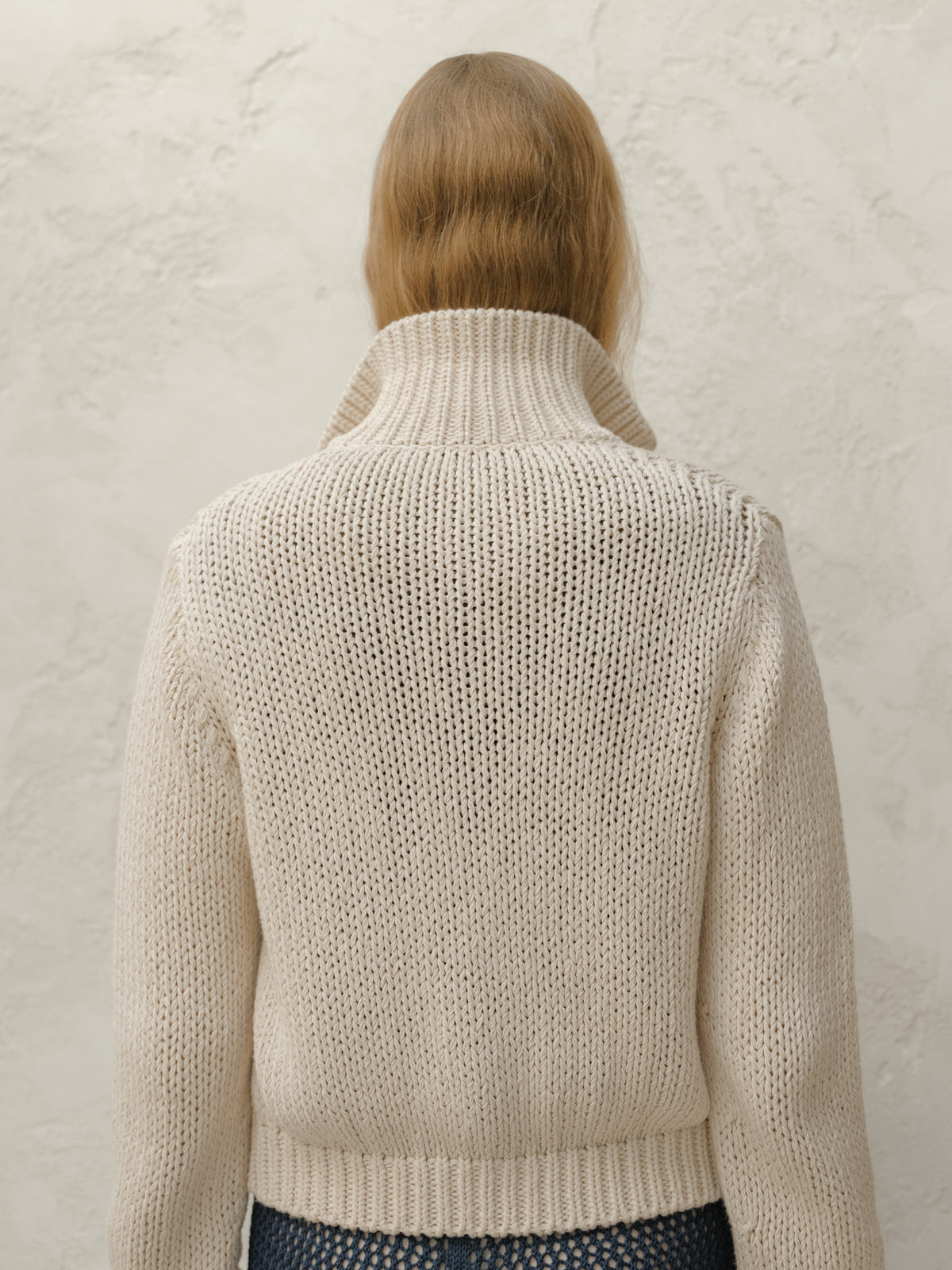 Back view of a modella con a crema chunky knit bomber jacket con a orlo a coste against a texturosso light wall.