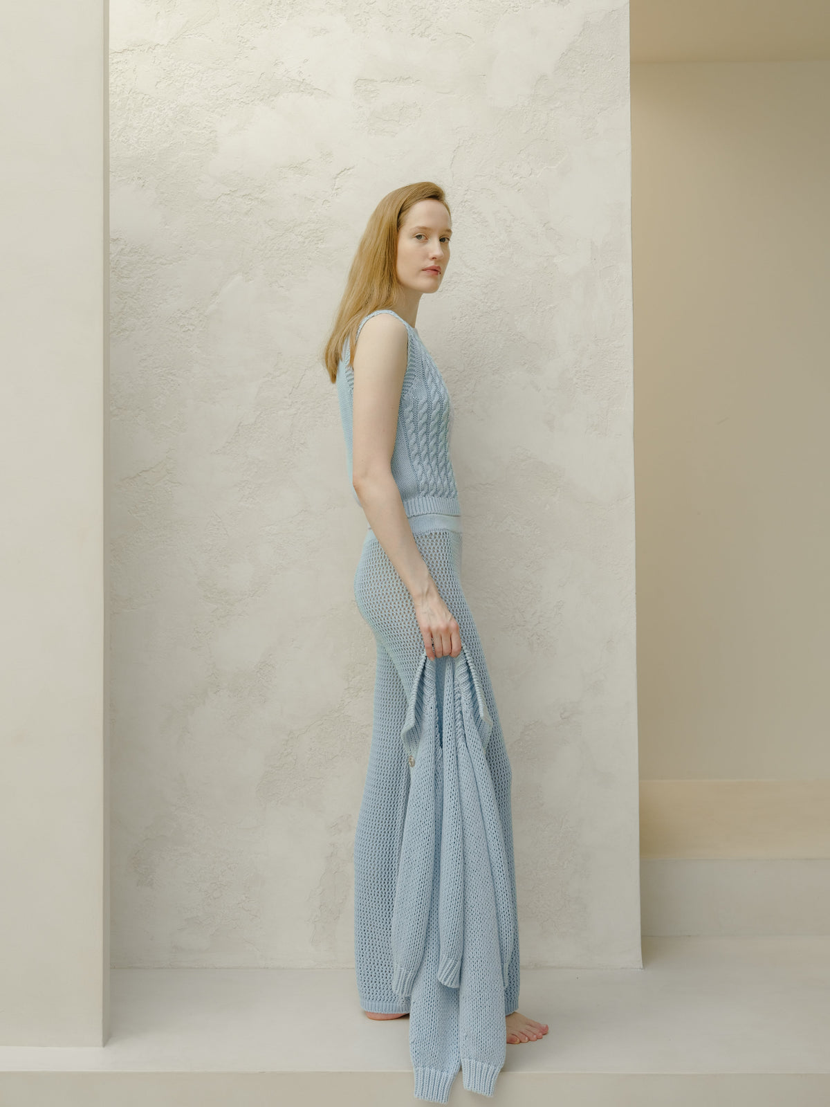Full-length vista laterale of a donna con a azzurro top in maglia senza maniche e matching mesh-knit pants against a pale texturosso backdrop.