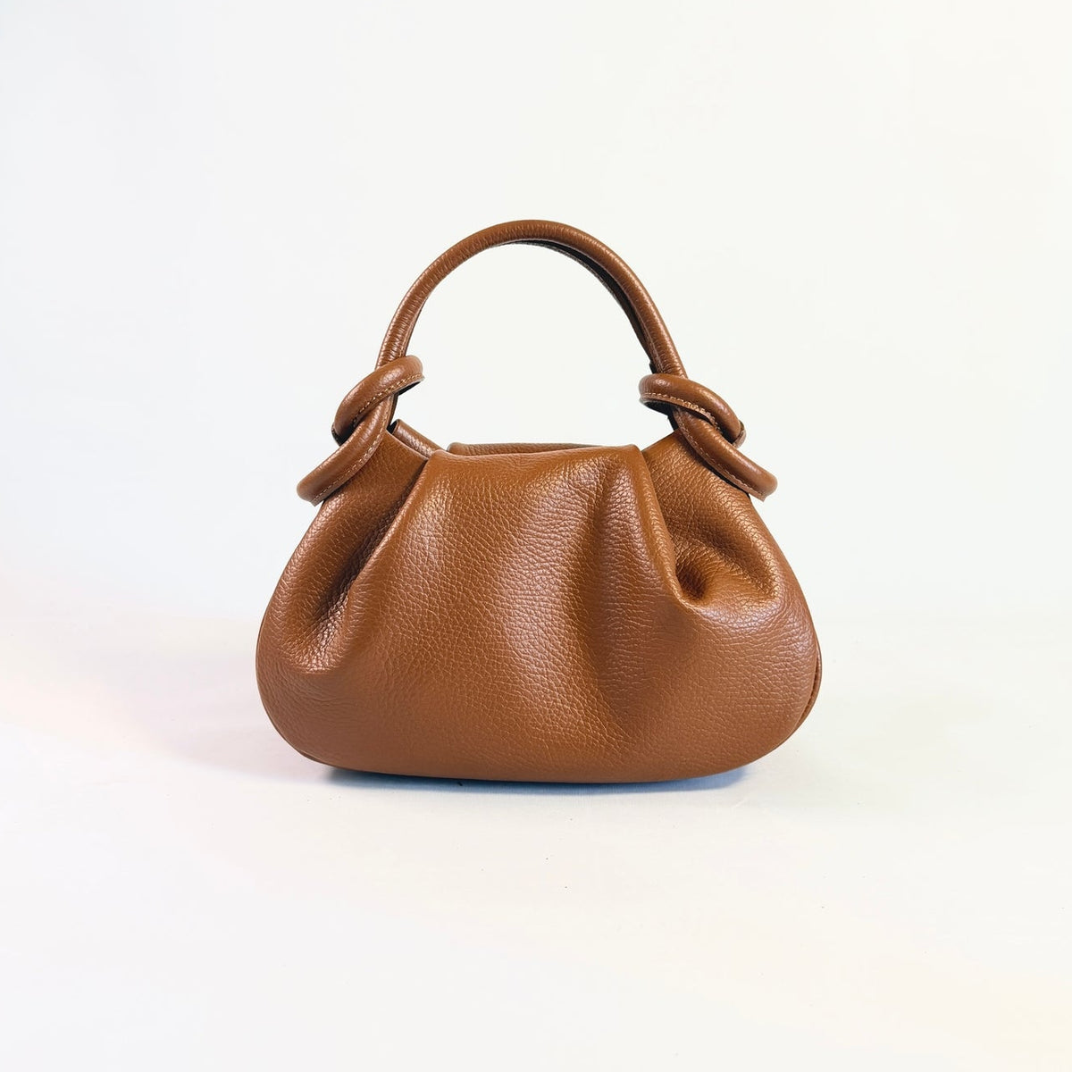Pequeño bolso Anello de piel granulada color coñac con asa superior y detalles de lazo sobre fondo blanco.