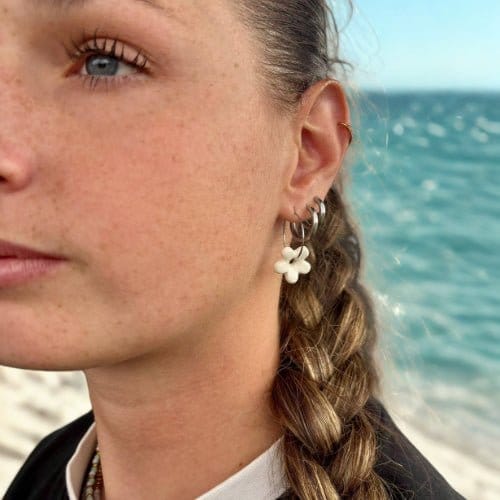 Pendiente colgante Mimì de cerámica blanca floral en la oreja izquierda de una mujer con el mar al fondo