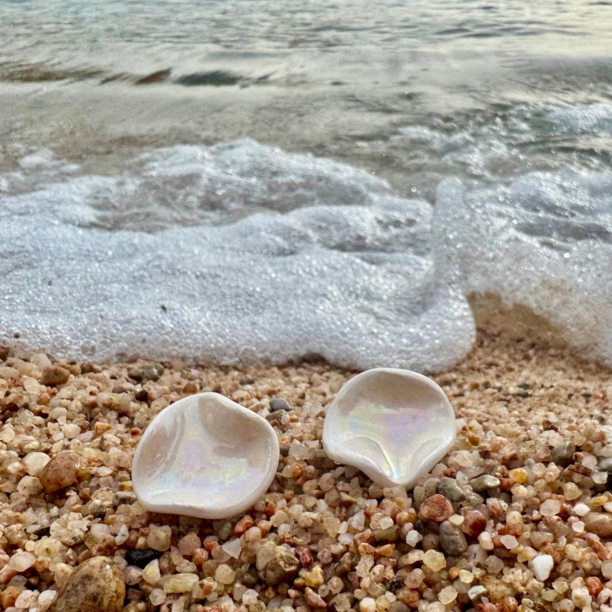 Due orecchini iridescenti a forma di conchiglia su una spiaggia di ciottoli con onde sullo sfondo