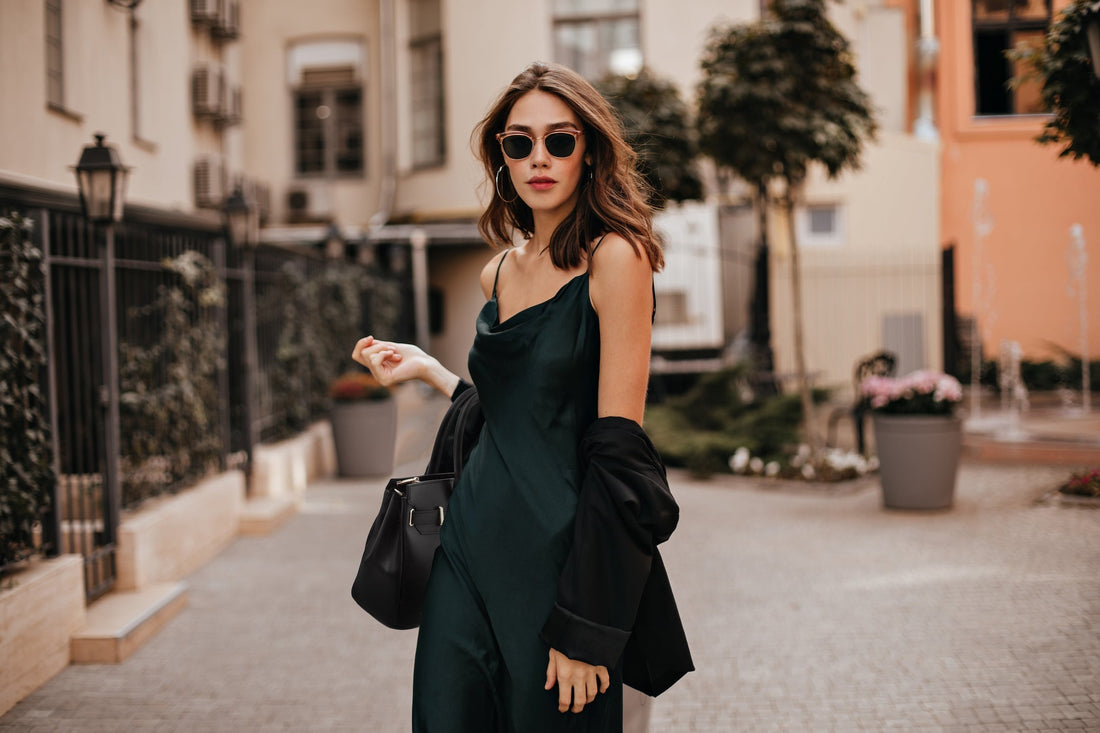 Stylish woman in a dark green slip dress and sunglasses walks down a city street carrying a black leather handbag.
