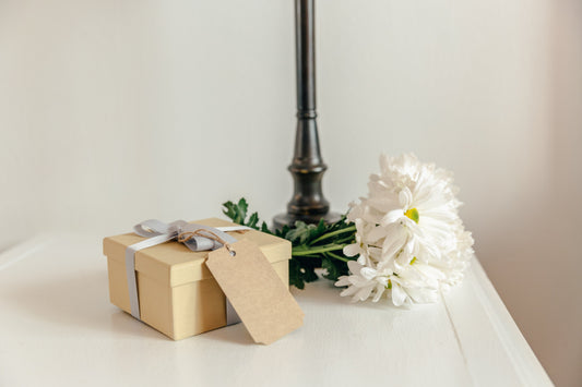 Beige geschenkdoos met grijs lint en blanco label naast een wit boeket op een wit oppervlak
