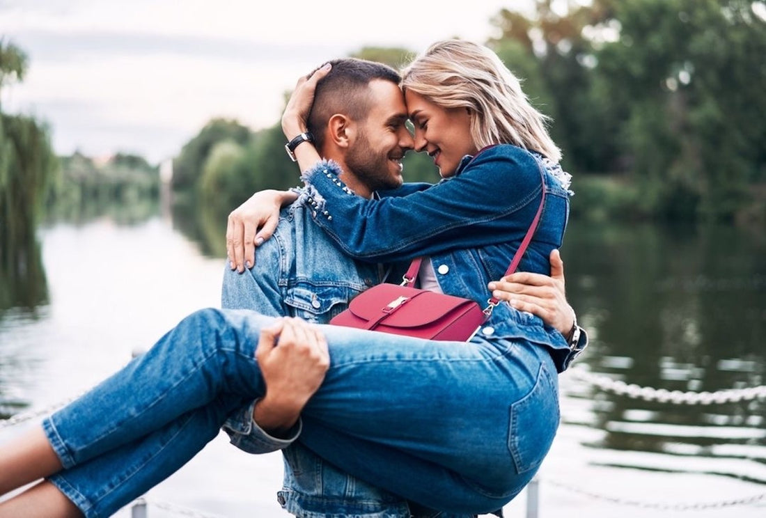Een romantisch stel in denim omhelst elkaar bij een meer met een felroze handtas.