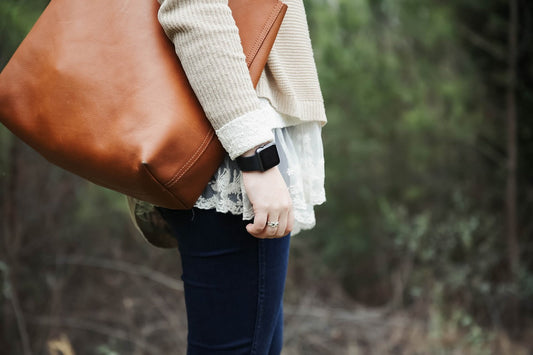Hellbraune Ledertasche über der Schulter einer Person in cremefarbenem Strickoberteil und Spitzenrock, im Freien zwischen Bäumen.