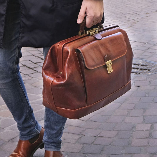 Person in Jeans trägt eine braune Lederaktentasche mit messingfarbenen Metallteilen durch eine Kopfsteinpflasterstrasse.