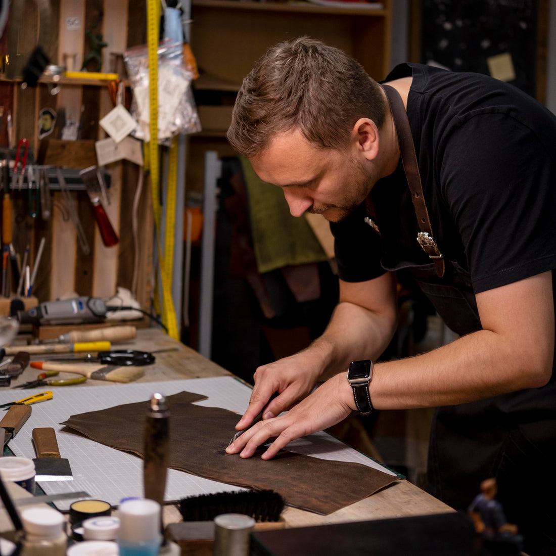 Leather craftsman cuts a dark hide on a cluttered workbench in a busy workshop.