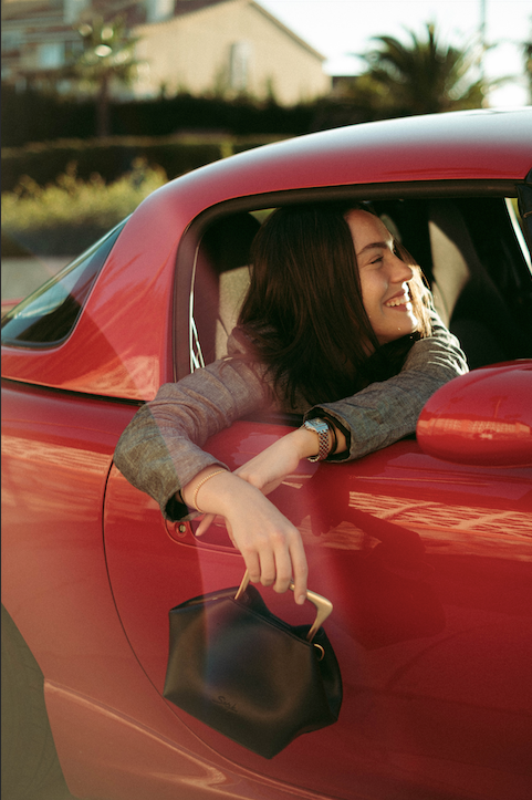 Smiling woman leaning from a red vintage car window while holding a small dark brown leather shoulder bag.