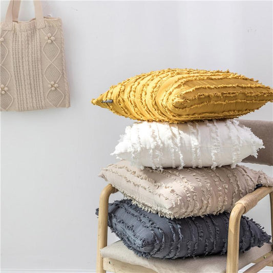 Gestapelde kussens met textuur in beige, wit en mosterdgeel op een houten stoel, met een geweven tas aan een witte muur.