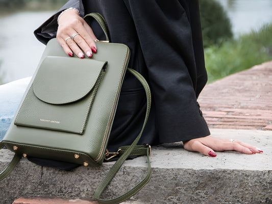 Grüne Lederhandtasche mit Vordertasche und goldfarbenen Metallteilen, neben einer Person in einem schwarzen Mantel gehalten.