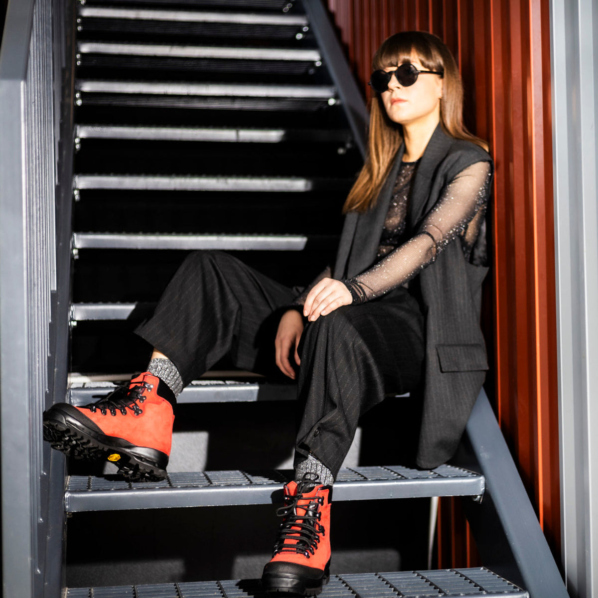 Woman in a dark suit with sheer sleeves sits on metal stairs wearing red hiking boots.
