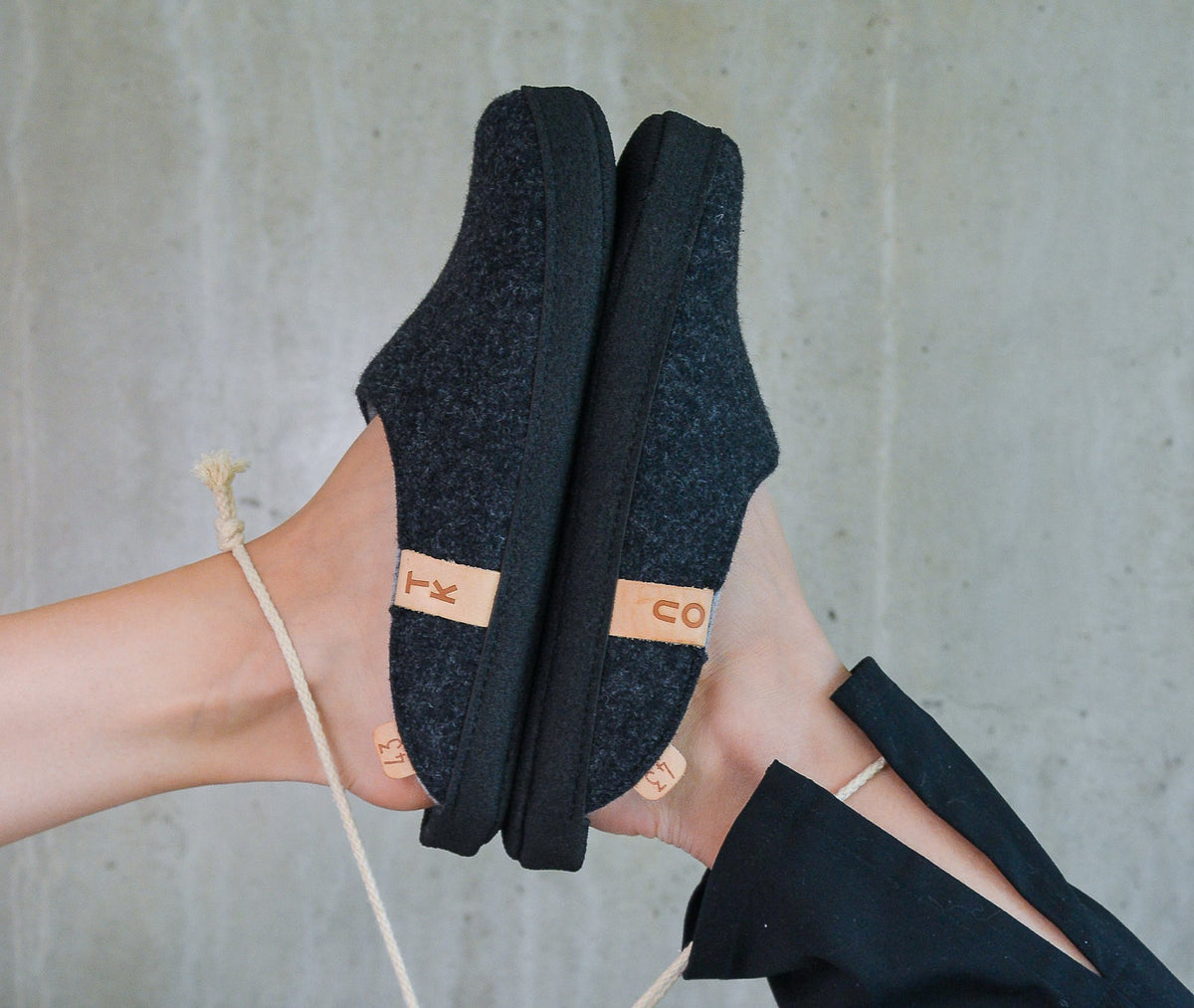 Bare feet wear black knit slippers with tan branding straps against a gray concrete wall
