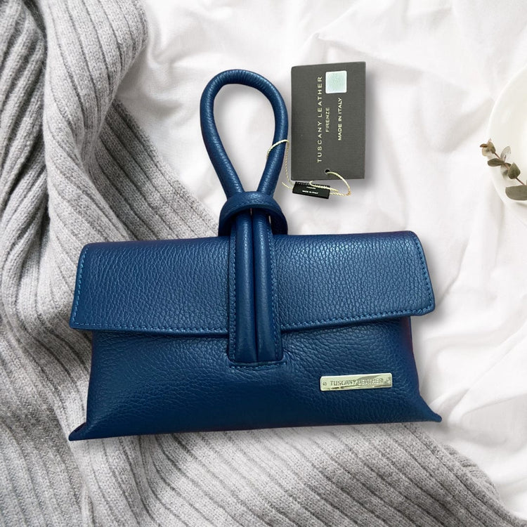 Blauwe TL Bag van Tuscany Leather op zachte grijze gebreide stof met zichtbaar productlabel, een elegant Italiaans leren accessoire.