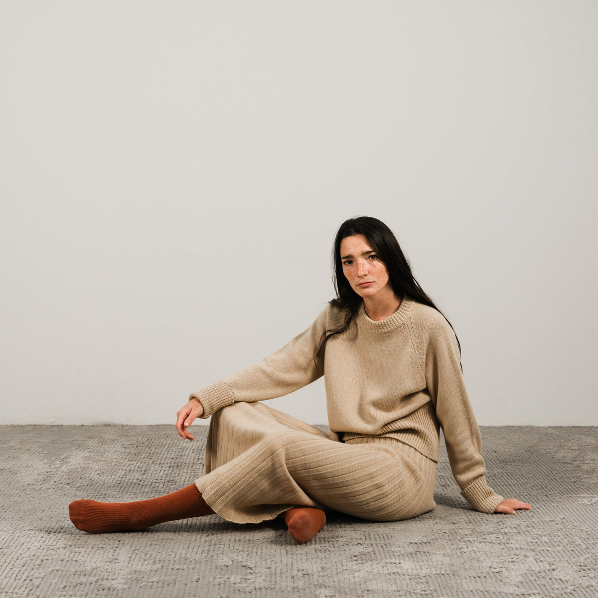 Woman wearing a beige cashmere knit set sits on a textured rug against a light neutral wall.