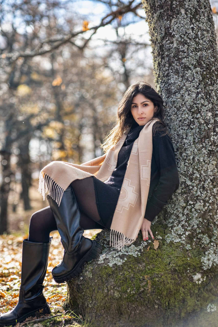 Femme portant une écharpe beige en laine d’alpaga, assise contre un arbre dans une forêt automnale paisible, avec des bottes noires et de la maille.