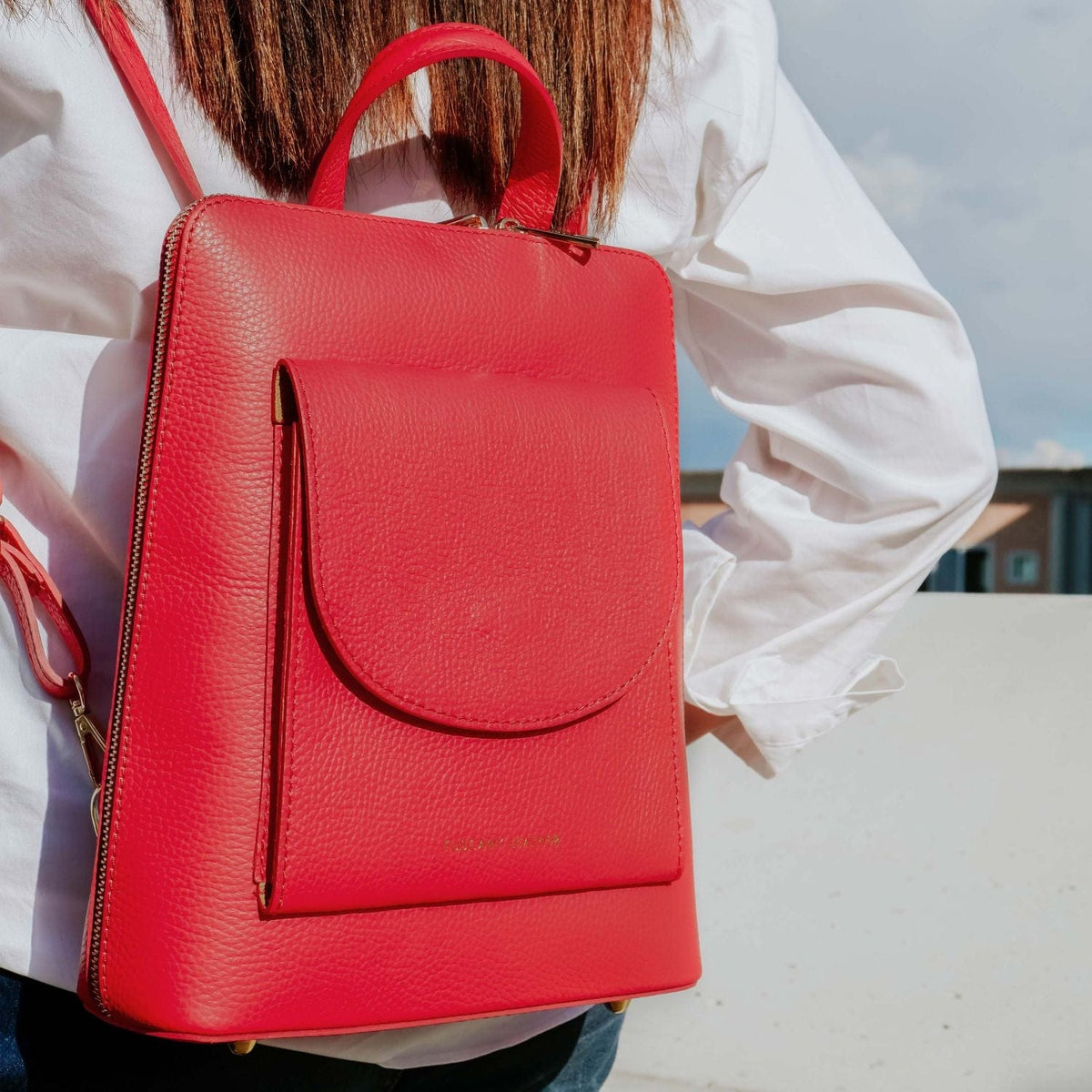 Leuchtend roter Lederrucksack mit gebogener Fronttasche und oberem Griff, getragen von einer Person im weissen Hemd.
