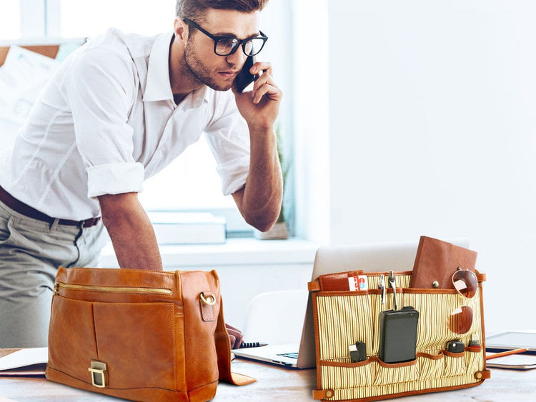 Mann in weissem Hemd und mit Brille telefoniert neben cognacfarbenen Leder-Businesstaschen und einem Laptop auf einem Schreibtisch.