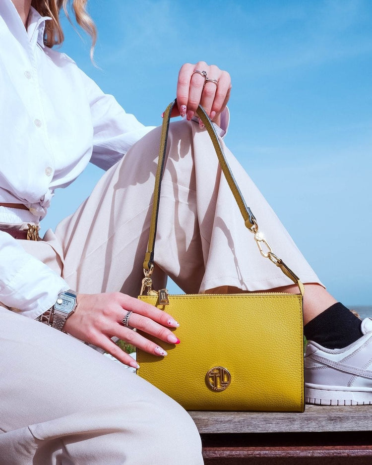 Bright yellow leather clutch with a top handle and gold hardware held by a person in a white shirt.