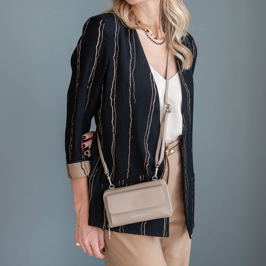 A woman models a taupe leather crossbody wallet bag with a silver-tone logo plate. She is wearing a black and beige striped blazer, a white top, and beige trousers against a solid slate gray background.
