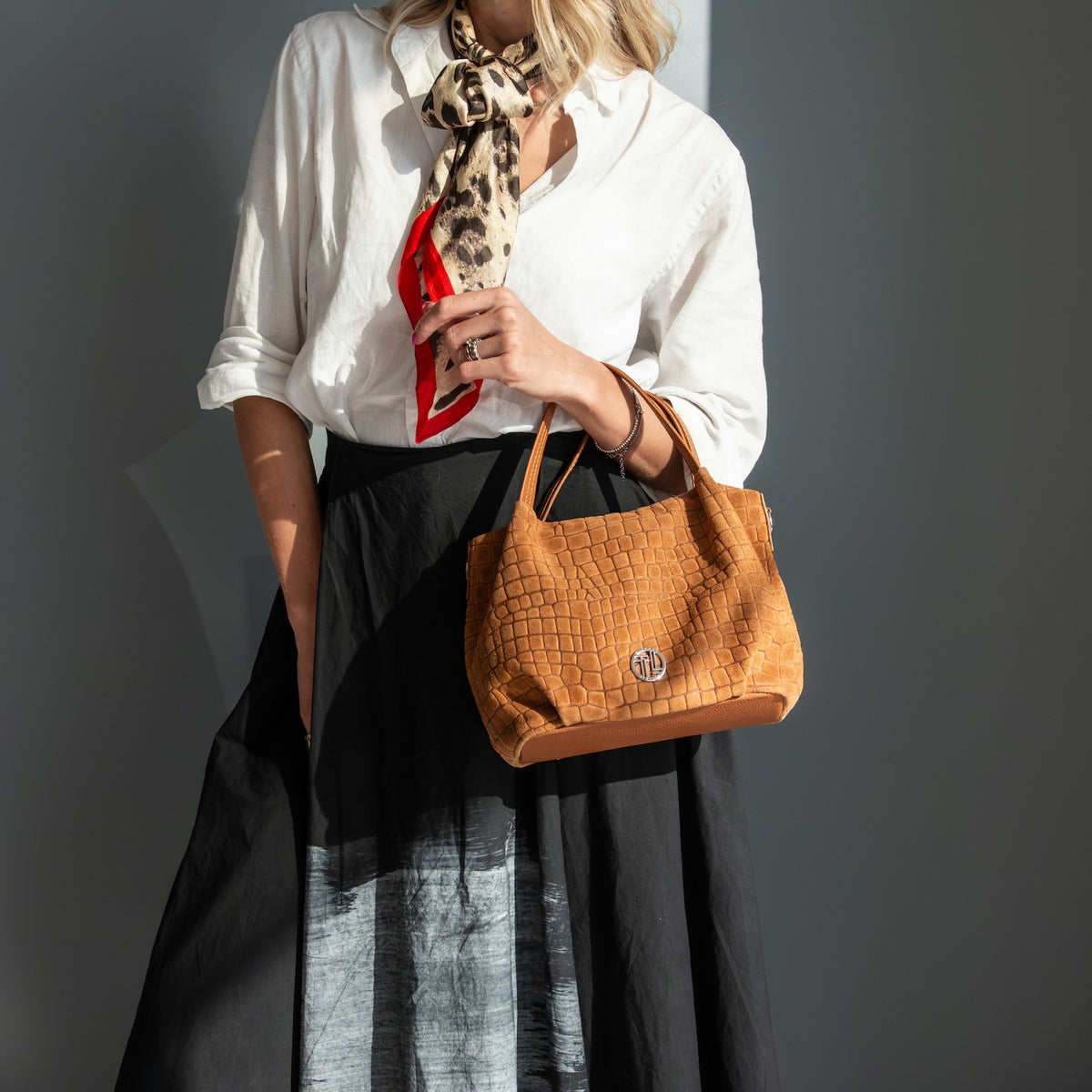 A woman wearing a white blouse, black skirt, and a leopard-print neck scarf holds a tan crocodile-embossed suede leather handbag with a silver-tone logo on a dark gray background.