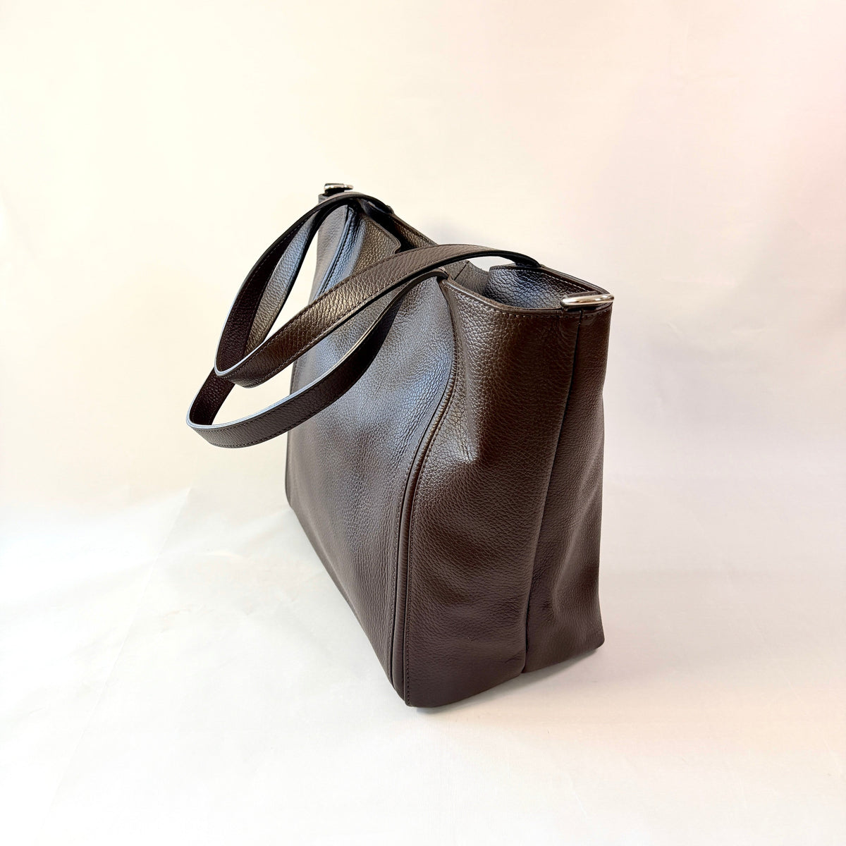 A dark brown pebbled leather tote bag with a single shoulder strap, shown from a side angle on a plain white background.
