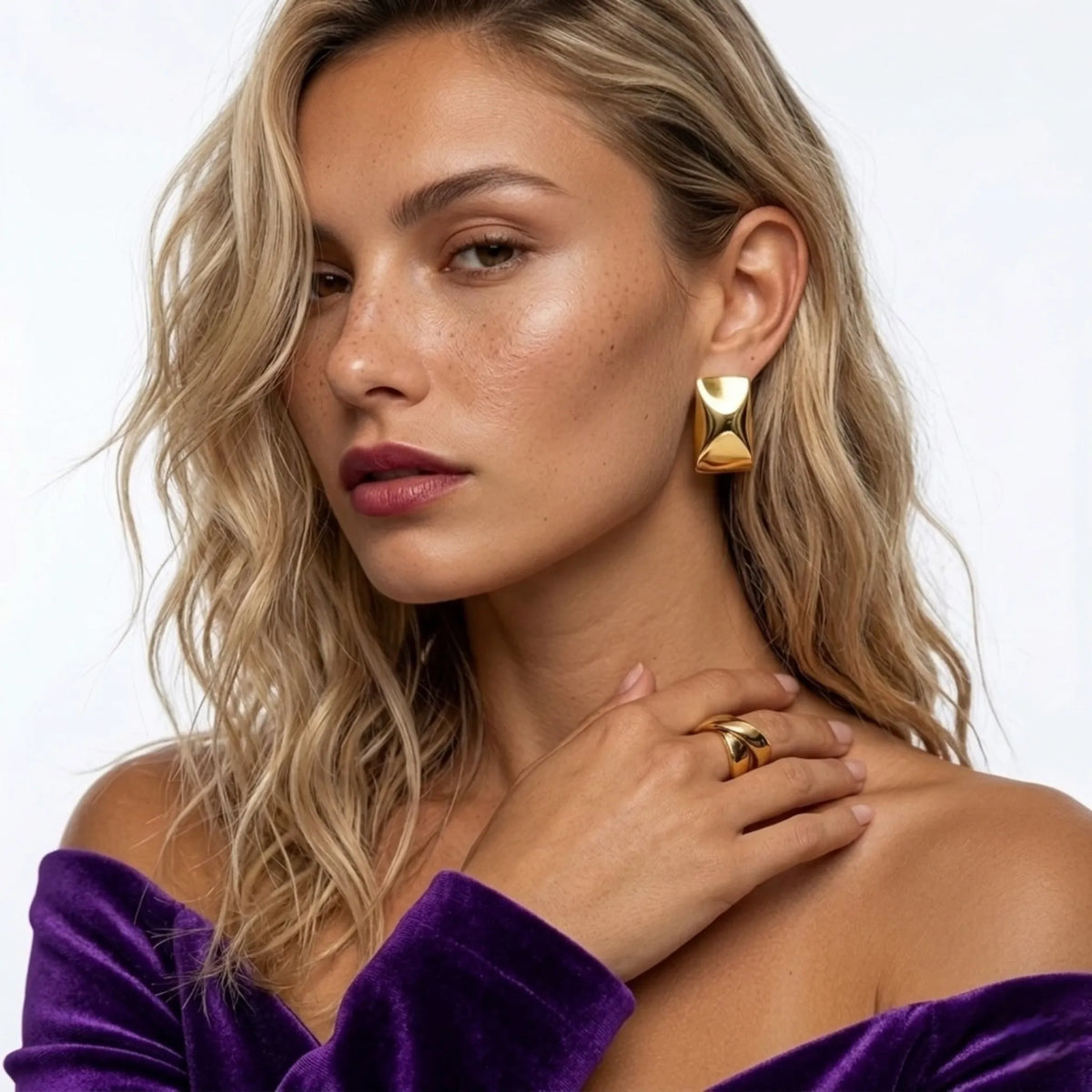 Frau trägt gold earrings and ring, with a purple top against a white background