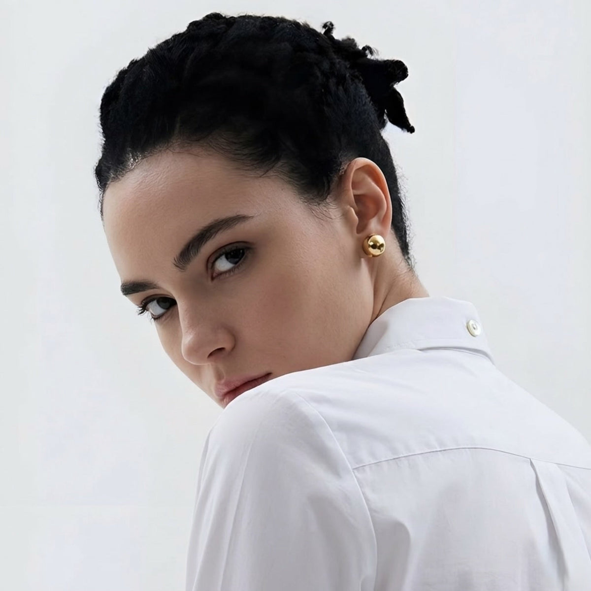 Woman with braided hair wearing gold earrings and a white shirt against a light background