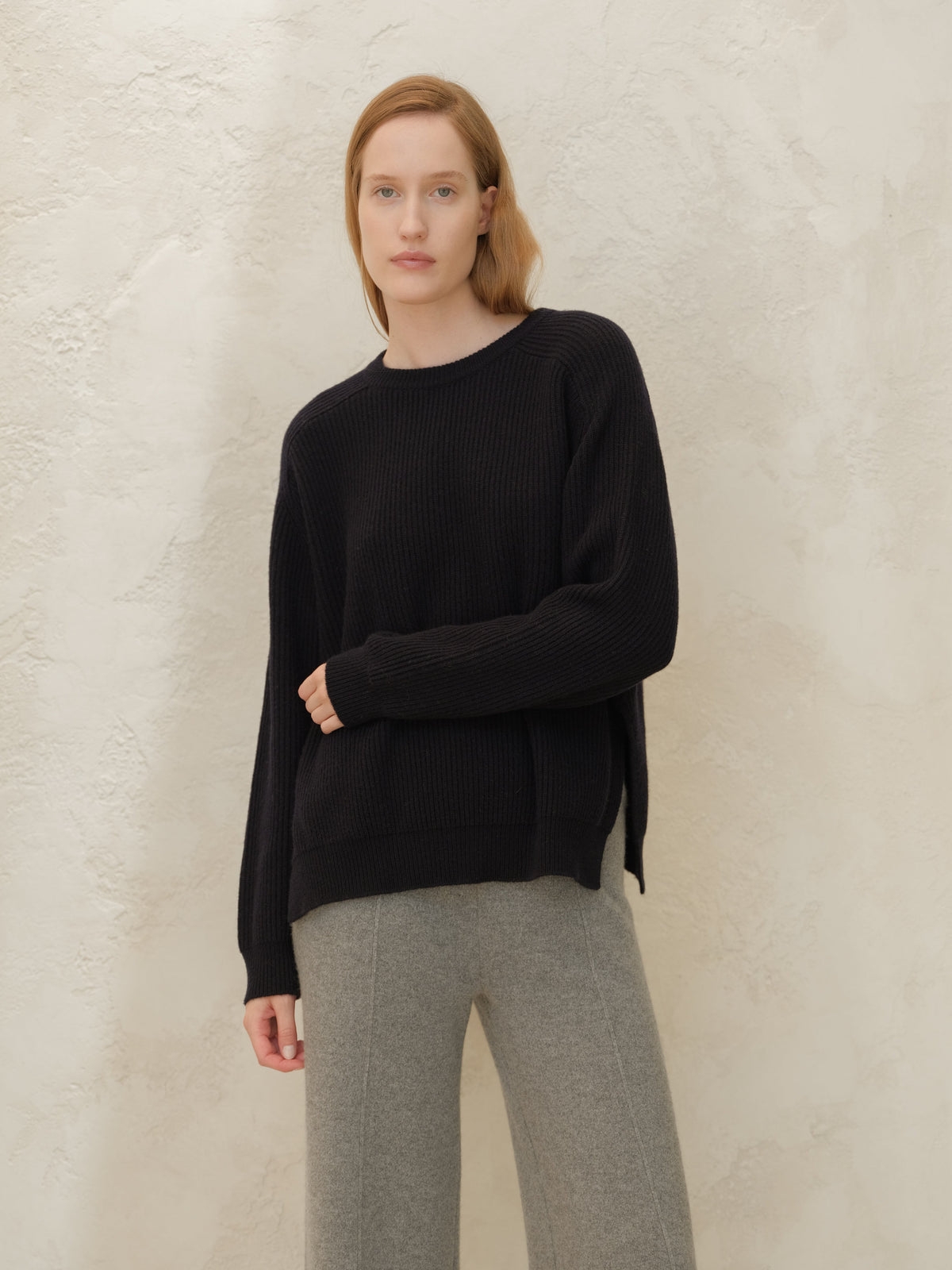 Modelo con black cashmere mix crewneck ribbed sweater with side slits and gray pants against a textured wall