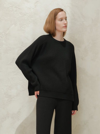 Modelo con black cashmere mix crewneck ribbed sweater with side slits against a textured light wall