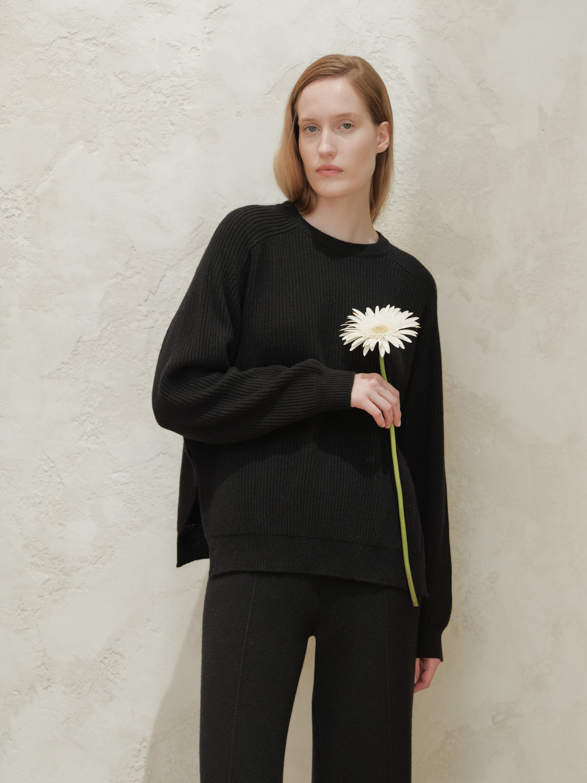 Modelo con a black ribbed cashmere mix jersey de cuello redondo with side slits holding a white flower against a textured beige wall.