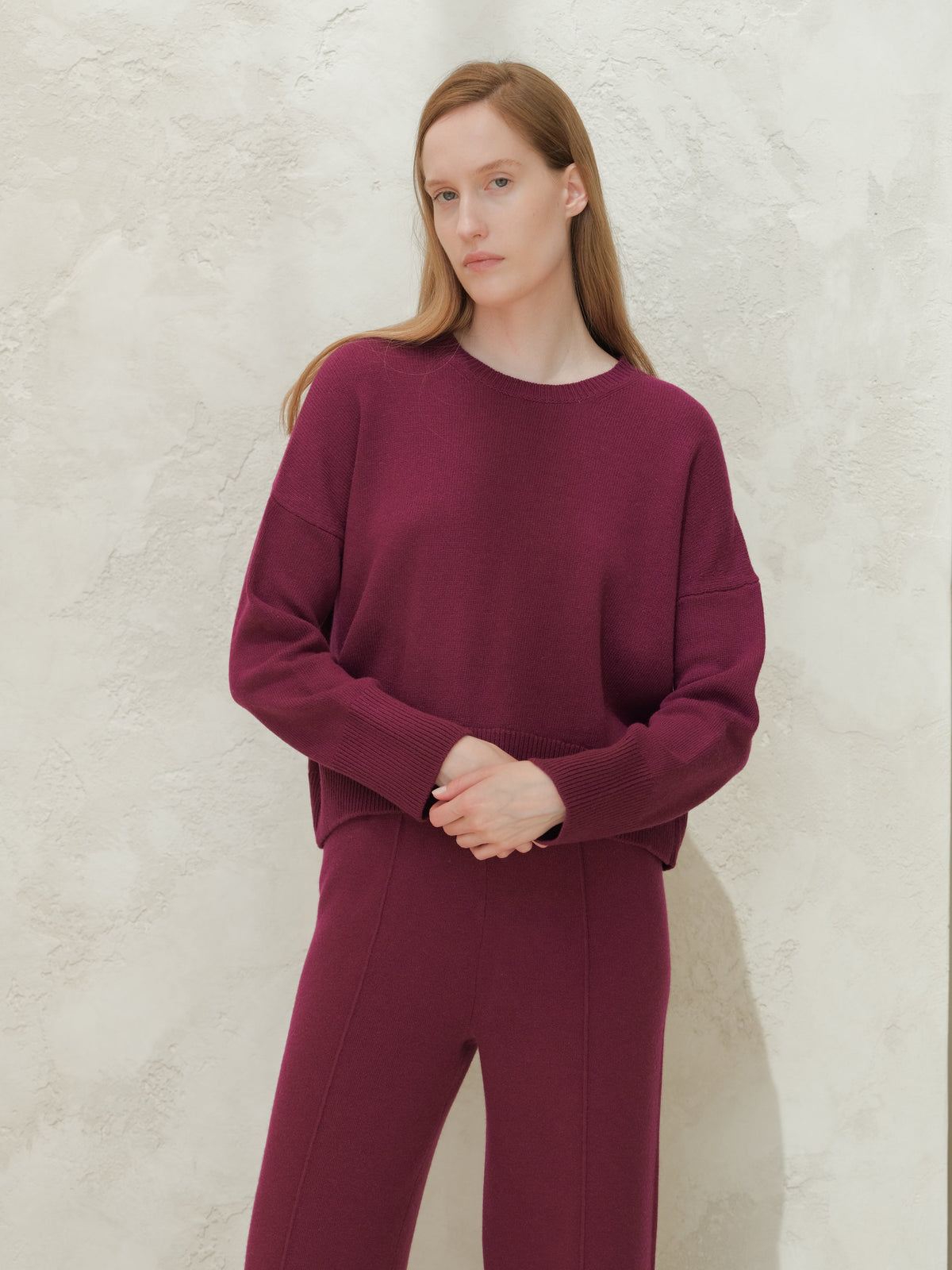 Model wearing a deep burgundy cashmere mix crewneck sweater with matching knit pants against a textured light wall