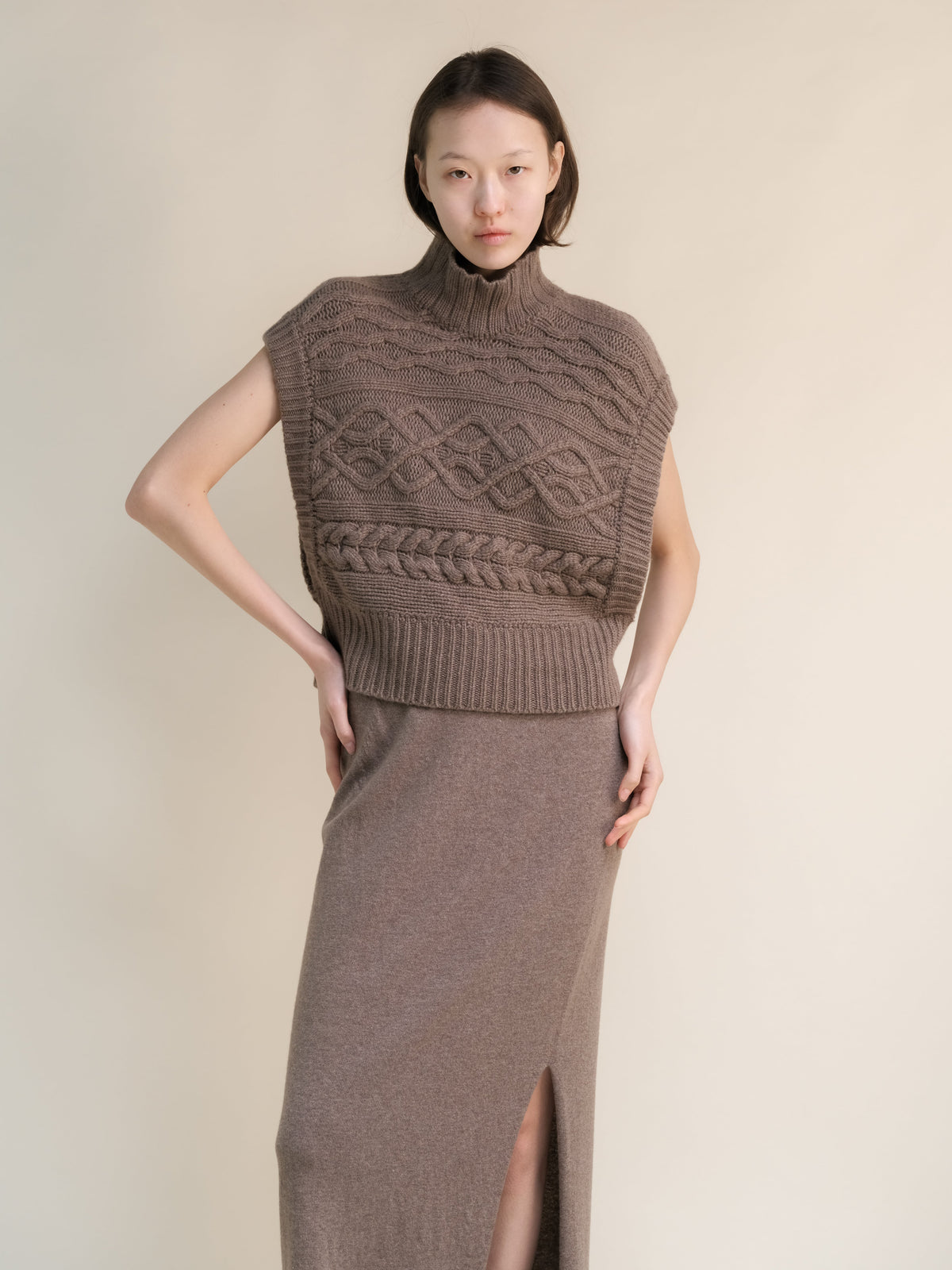 Taupe cashmere mix maxi cable tunic styled as a sleeveless high neck poncho with intricate knit patterns and side openings.