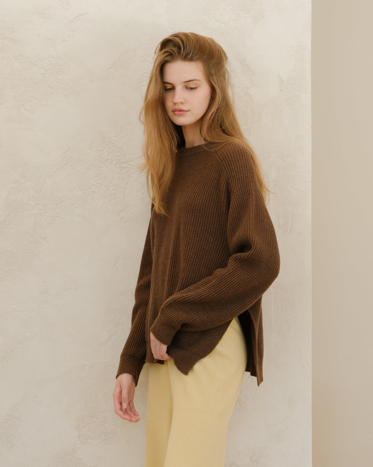 Modelo con a brown ribbed knit jersey de cuello redondo with side slits, paired with beige pants, standing against a textured wall.