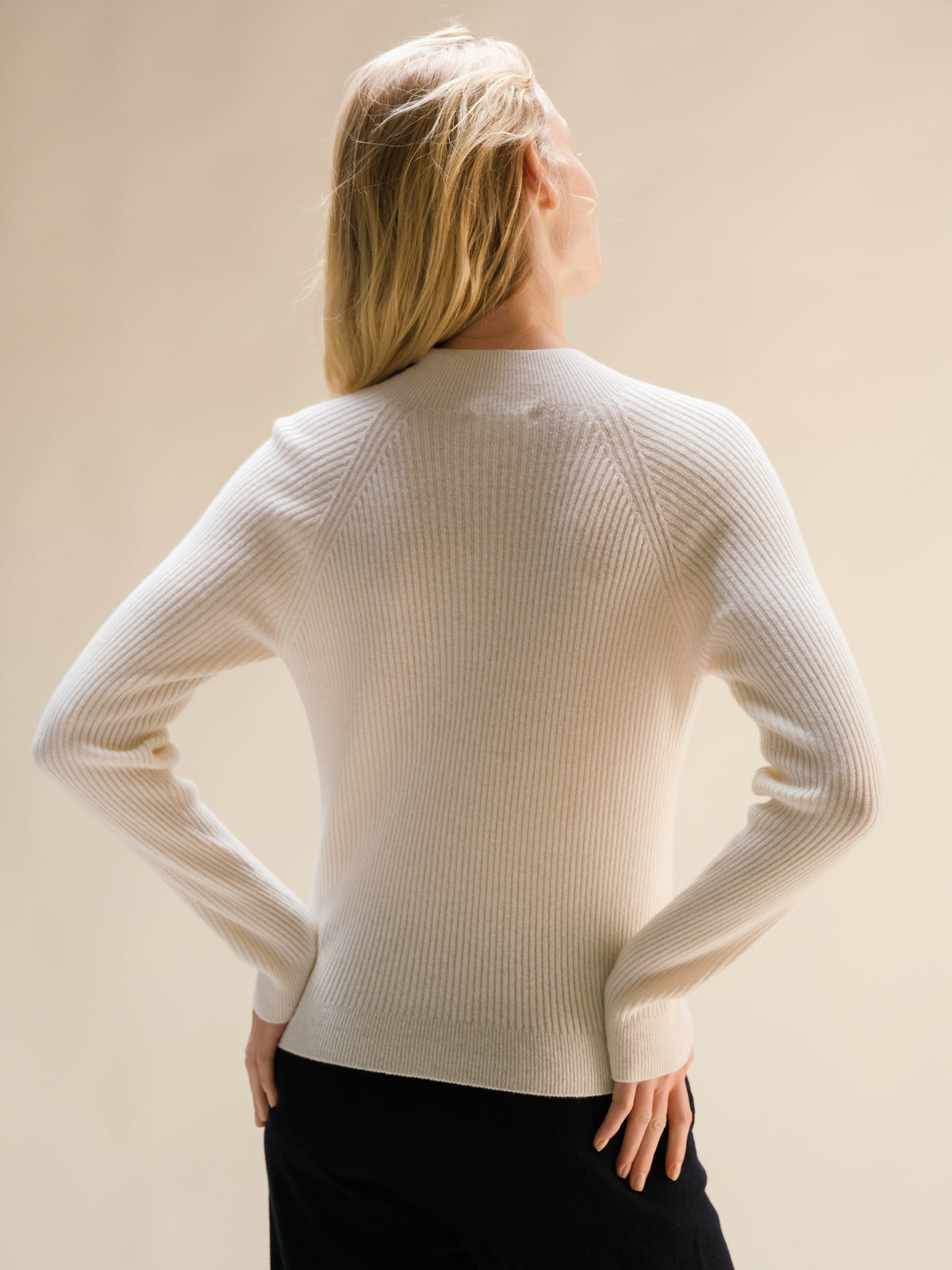 Model wearing cream ribbed cashmere mix mock neck sweater, back view with hands on hips.