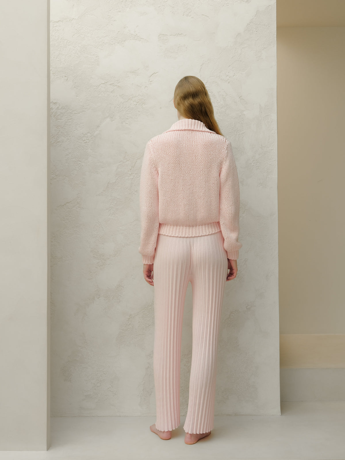 Back view of a modella con a pale rosa knit sweater e matching ribbed pants davanti a una parete testurizzata