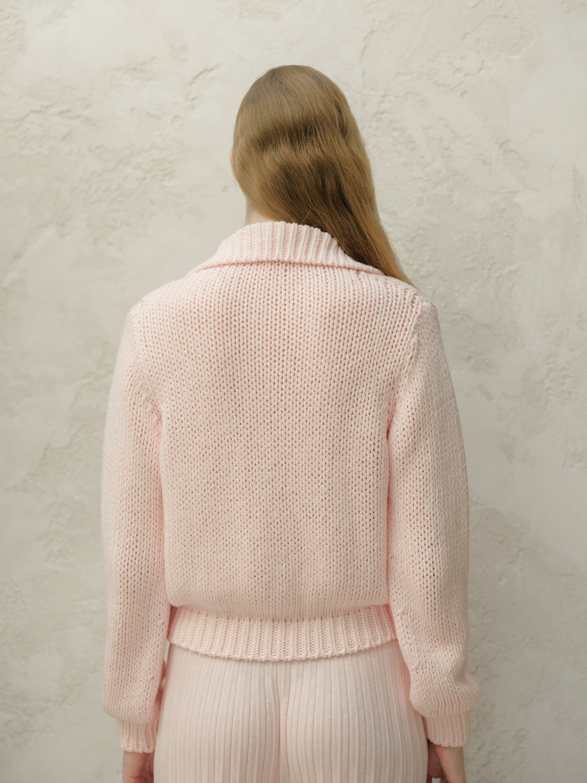 Back view of a model in a rosa tenue knit cardigan showing vestibilità rilassata e orlo a coste davanti a una parete testurizzata