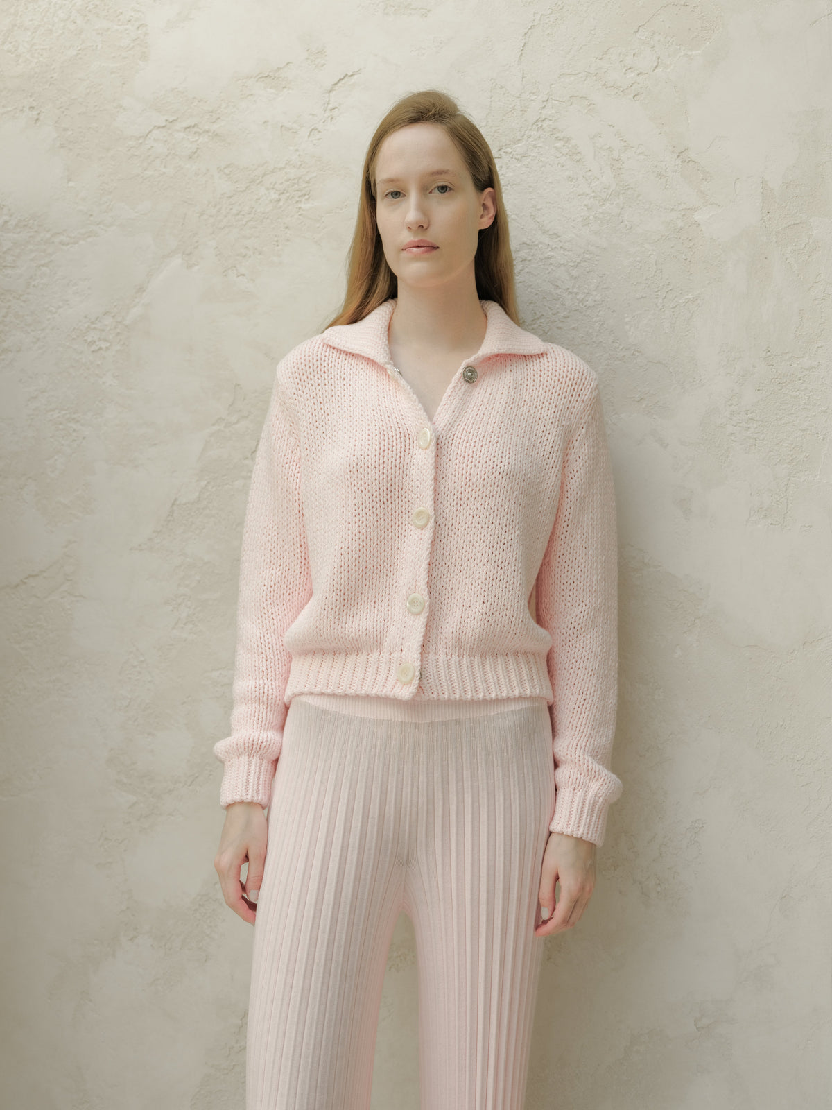 Vista frontale of a modella con a rosa tenue knitted cardigan con large bianco buttons against a texturosso beige wall